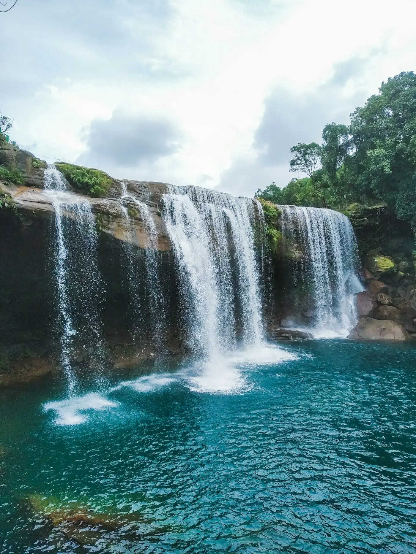 A wide curtain waterfall pouring into a turquoise pool near Shillong